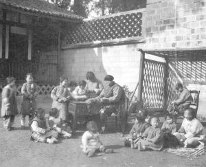 街角玩耍的小孩。 他们可能是慈善堂收养的小孩，在慈善堂工人的照看下，他们在冬日的阳光下嬉戏。照片显示了成都典型的高墙院落。那爱德摄，1910年。